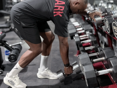 Ilias Skali en tenue Gymshark, penché près d’un rack d’haltères en salle de musculation.