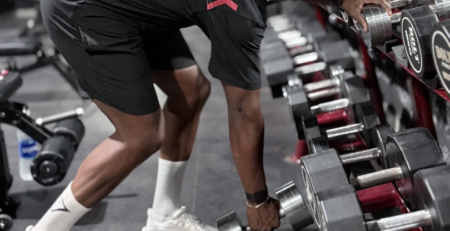 Ilias Skali en tenue Gymshark, penché près d’un rack d’haltères en salle de musculation.