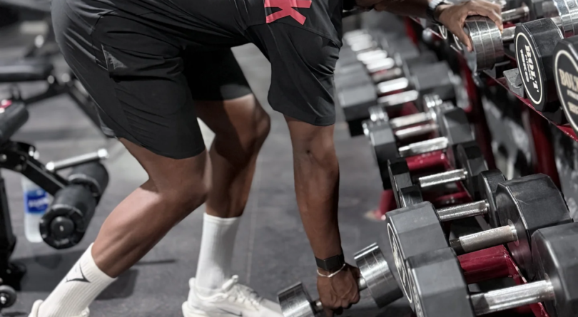 Ilias Skali en tenue Gymshark, penché près d’un rack d’haltères en salle de musculation.